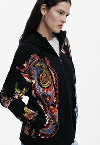 Svart zip-up hoodie med färgglada abstrakta mönster på ärmarna, tillverkad av mjukt tyg med en slät yta. Huvdesign med fickor.