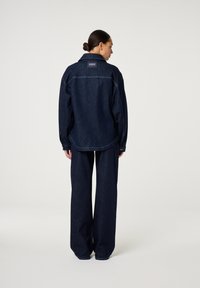 Spijkerjack en wijde broek in donkerblauw. Het jack heeft een achterkant met schouderstuk, zijzakken en een logopatch; de broek heeft een hoge taille en een losse pasvorm.