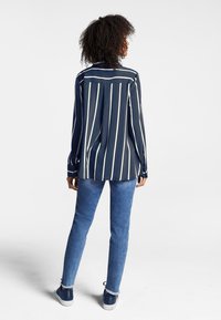 Mujer con cabello rizado de pie, de espaldas, vestida con una camiseta de rayas azul marino y blanca de manga larga, jeans azules y zapatillas azul marino.