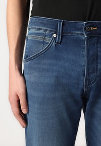Jean en denim bleu foncé avec une texture lisse, comportant cinq poches, un zip à bouton et des détails de couture contrastés.