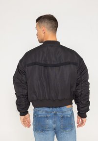 Czarna kurtka bomber z kieszenią z zamkiem błyskawicznym na plecach, prążkowanymi mankietami i elastycznym dołem. Gładki materiał z wyściełanym, luźnym krojem.