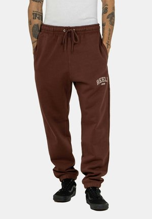 Pantalones de chándal marrones con puños elásticos, cintura con cordón ajustable y logo bordado en blanco "REEL" en la pierna izquierda. Textura de tela suave.