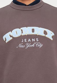 Šedá mikina s okrúhlym výstrihom má textúrované svetlomodré logo "TOMMY" a biele texty "JEANS New York City" nižšie. Hladká textúra látky.