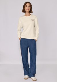 Felpa crema con il testo "CITY CLUB", abbinata a pantaloni di jeans blu larghi con strisce laterali rosse e sneakers bianche.