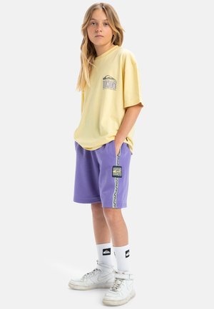 Geel T-shirt met korte mouwen en logo, gecombineerd met paarse shorts met een logo-patch aan de zijkant en een patroonrand. Witte high-top sneakers.