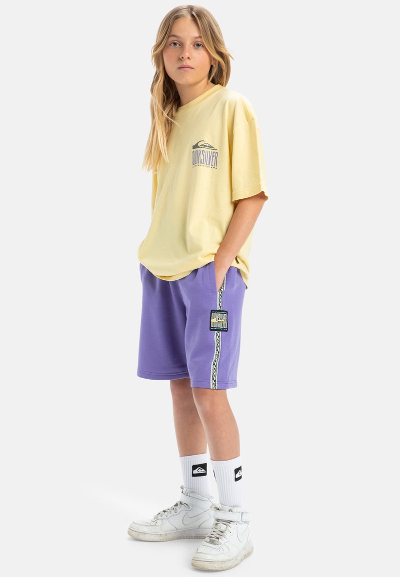 Geel T-shirt met korte mouwen en logo, gecombineerd met paarse shorts met een logo-patch aan de zijkant en een patroonrand. Witte high-top sneakers.