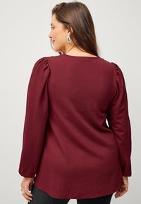 Burgunderfarbener, strukturierter Oberteil mit langen Ärmeln und gerafftem Detail an den Schultern, ausgestattet mit einem runden Ausschnitt und lockerer Passform, kombiniert mit schwarzen Leggings.