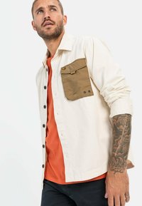 Knoopshirt in crème met een bruine borstzak; oranje onderhemd zichtbaar eronder. Beschikt over lange mouwen en een relaxte pasvorm.