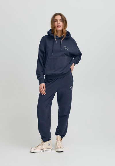 Felpa blu navy con tasca a marsupio e cordini, abbinata a pantaloni joggers coordinati. Il materiale appare morbido e dalla texture piacevole. Accenti del logo in bianco.