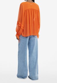 Top in chiffon arancione con pieghe, maniche lunghe e dettagli con lacci; abbinato a jeans in denim azzurro a gamba larga.