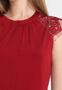 Femme portant un haut sans manches rouge avec des détails en dentelle sur les épaules et un petit plissé à l'avant près du col.
