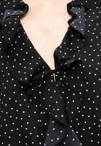 Μαύρο μπλουζάκι με λευκά πουά, διαθέτει βαθύ V-λαιμόκοψη, κορδόνι δέσιμο, γιακά με βολάν και ορατή λεπτομέρεια με κουμπιά στο στήθος.