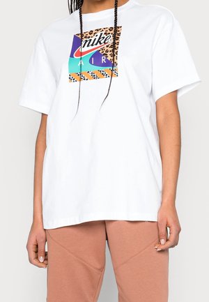 Personne portant un T-shirt Nike Air blanc surdimensionné avec un motif de patch léopard coloré et rayé, associé à un pantalon marron clair.