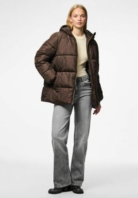 Chaqueta acolchada marrón con capucha, que presenta un cierre de cremallera, bolsillos laterales y una textura suave similar al satén, combinada con jeans grises de tiro alto.