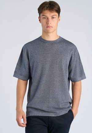 Jonge man met kort bruin haar, gekleed in een loszittend grijs T-shirt met korte mouwen en donkere broek, staand tegen een effen lichte achtergrond.
