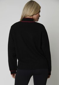 Donna con capelli biondi che indossa un maglione nero con colletto e polsini a righe bordeaux e neri, rivolta di spalle su uno sfondo neutro.