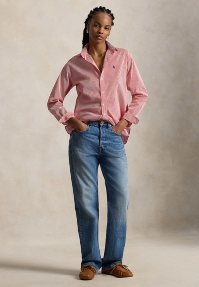 Γυναίκα με πλεξούδες, φοράει πουκάμισο ροζ με κουμπιά, μπλε τζιν και καφέ μοκασίνια, στέκεται με τα χέρια στις τσέπες μπροστά σε απλό φόντο.