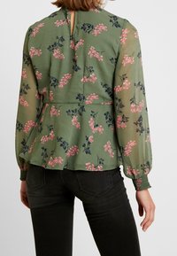 Floral peplum blouse in olive green with sheer long sleeves, pink and navy floral pattern, back keyhole opening, and gathered cuffs.