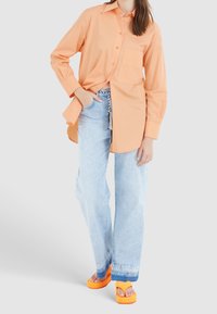 Chemise boutonnée orange clair à manches longues avec une poche poitrine, associée à un jean bleu clair ample et des sandales à plateforme orange vif.