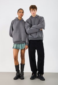 Sudaderas grises de gran tamaño con bolsillos frontales; un modelo las combina con una falda de mezclilla con pliegues y botas de caña alta negras, el otro con pantalones negros y zapatillas.