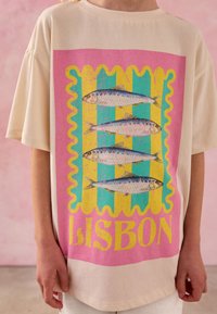 Krém színű bő fazonú póló, amely rózsaszín, türkiz és sárga grafikai mintát ábrázol szardíniákkal és "LISBON" felirattal. Puha pamut anyagból készült.