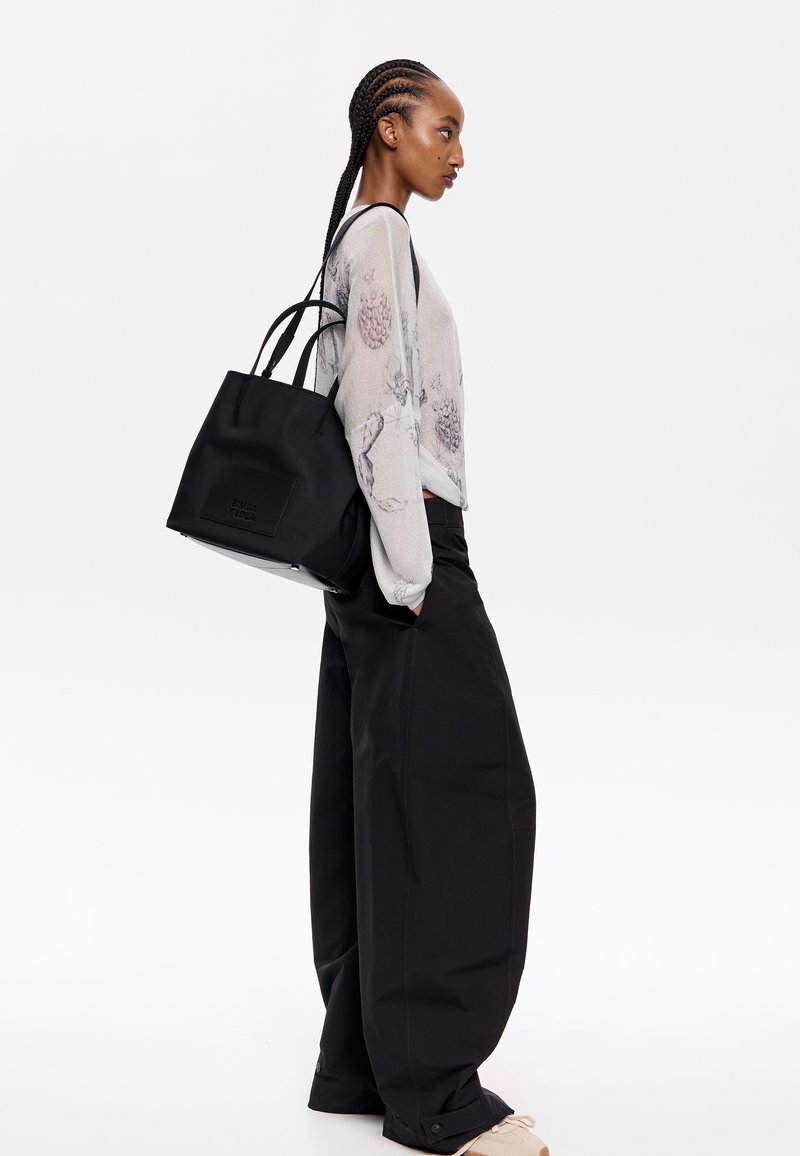 Borsa a tracolla nera con esterno testurizzato e un logo sottile. Indossata a spalla, abbinata a pantaloni neri oversize e una blusa leggera e a fantasia.
