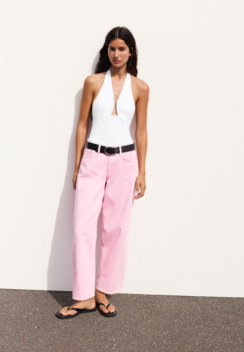 Femme aux longs cheveux foncés portant un haut dos-nu blanc, un pantalon rose à jambes larges, une ceinture noire et des tongs noires, debout contre un mur blanc.