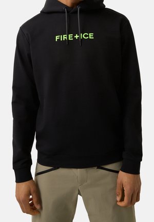 Czarna bluza z kapturem z neonowo zielonym napisem "FIRE + ICE" na piersi, noszona z jasnobeżowymi spodniami z poziomymi detalami zamków.