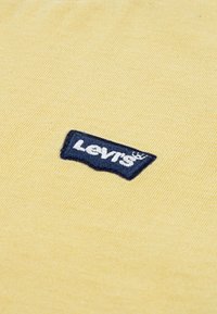 Κίτρινο βαμβακερό ύφασμα με μπλε κεντημένη ετικέτα λογότυπου Levi's, σε ορθογώνιο σχήμα και με υφή επιφάνειας.
