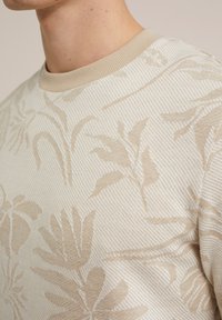 Närbild av en person som bär en beige och krämfärgad randig tröja med ett abstrakt botaniskt mönster och en beige ribbad rundhals.