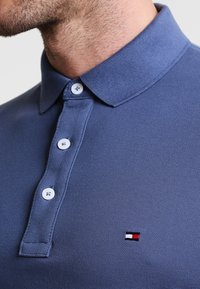 Modrá polo košile s texturovanou tkaninou. Obsahuje knoflíkovou légou se třemi knoflíky a malé vyšívané logo v červené, bílé a černé barvě na hrudi.