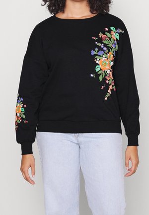 Black long-sleeve sweatshirt with colorful floral embroidery on chest and left sleeve, worn with light blue jeans.