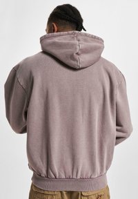 Hanorac cu glugă dintr-un material moale, de culoare mauve estompat. Are margini și manșete cu dungi, un croi lejer și cusături vizibile pe margini.