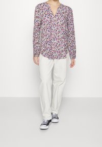 Langschedige, knoopsluiting blouse met een multicolore bloemenpatroon op een lichte achtergrond; gecombineerd met witte broeken en zwarte sneakers.