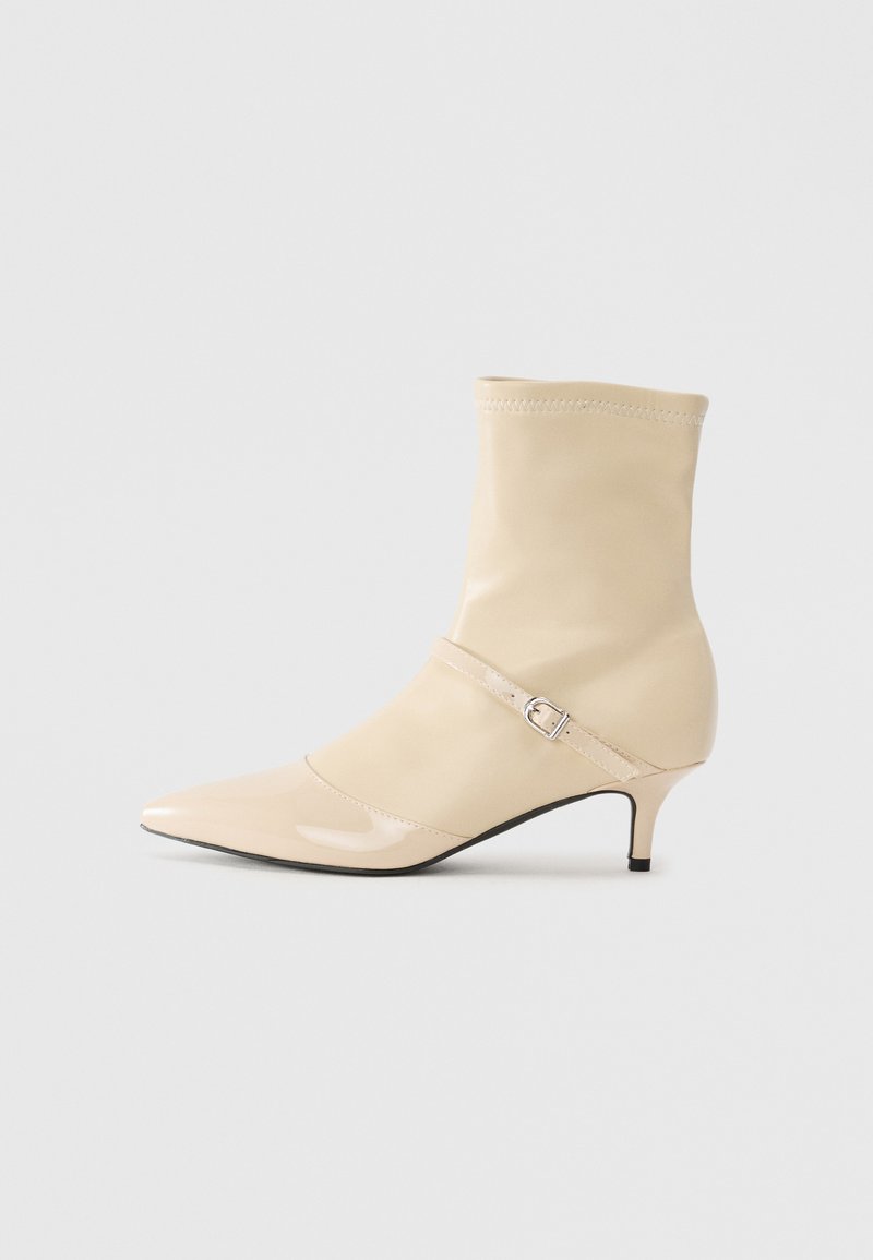 Beige mid-calf pointed toe ankle boot with a small kitten heel and decorative strap with silver buckle on a plain background.