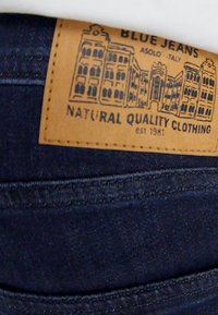 Niebieskie dżinsy denimowe z beżową skórzaną metką. Metka zawiera czarną ilustrację budynku oraz napis "Natural Quality Clothing."