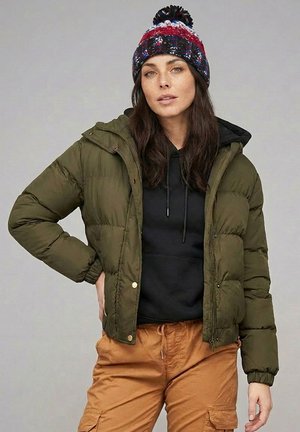 Woman wearing a multicolored knit hat, green puffer jacket, black hoodie, and tan cargo pants standing with one hand on her hip against plain background.