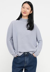 Žena s dugom crnom kosom koja nosi svijetloplavi turtleneck pulover i tamnoplave traperice, s rukama u džepovima, na bijeloj pozadini.