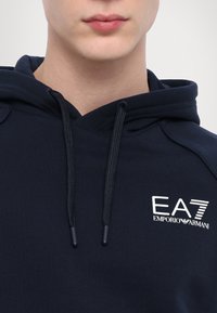 A tengerészkék kapucnis pulóver húzózsinórral és fehér hímzett EA7 Emporio Armani logóval rendelkezik a mellrészén. Puha anyag, sima textúrával.