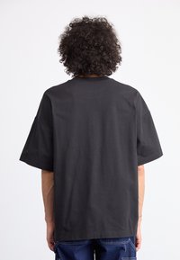 Person with curly hair wearing a plain black oversized T-shirt and dark blue pants, facing away against a white background.