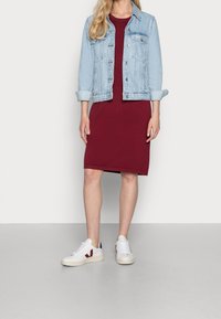Vrouw in een lichtblauwe spijkerjack, knielange bordeauxrode jurk en witte sneakers staand tegen een effen achtergrond.