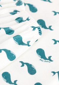 Tessuto in cotone con un motivo ripetuto di balene color turchese su sfondo bianco, con dettagli di elastico arricciato. Etichetta del marchio visibile.