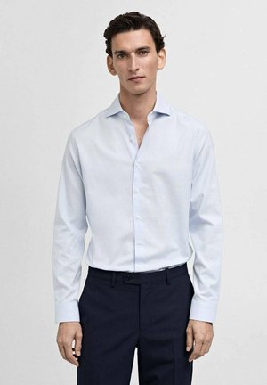 Cămașă bleu deschis, cu un model subtil, având guler clasic și mâneci lungi, purtată cu pantaloni negri închis.