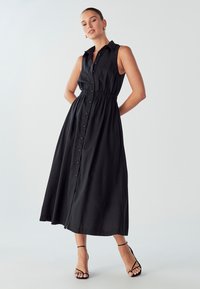 Juodas be rankovių suknelė su mygtukais, apykakle ir elastine juosimi, pasižyminti plaukiančiu siluetu ir minimalistiniu dizainu, derinama su dirželių sandalais.