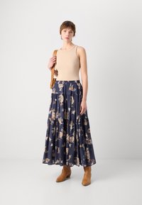 Lang, lagdelt marineblå skjørt med blomstermønster og en tettsittende beige tanktop. Brune ankelstøvler og en lys brun skulderveske fullfører antrekket.