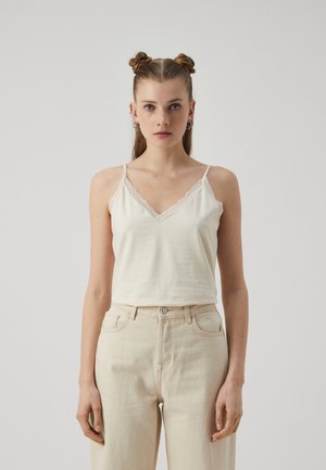 Lysebeige cami med blondekant, v-hals og tynne stropper, parret med høyhårede, lyse denimjeans.