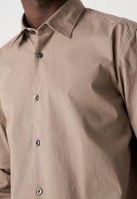 Hombre que lleva una camisa beige de botones con un cuello clásico, contra un fondo claro, recortada a la altura de la barbilla y la cintura.