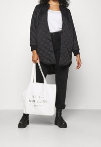 Persoon in zwarte gewatteerde jas, witte shirt, zwarte broek en zwarte laarzen, die een witte tote bag vasthoudt met de tekst "MADS NØRGAARD RECYCLED."