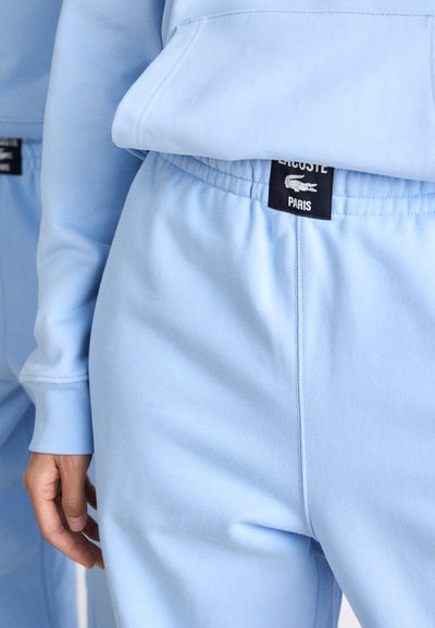 Sweatshirt bleu clair avec taille élastique, arborant une étiquette noire portant la mention "LACOSTE PARIS". Tissu lisse et coupe décontractée.