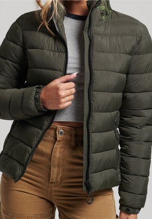 Woman wearing an olive green quilted jacket unzipped over a grey top, paired with high-waisted brown pants.